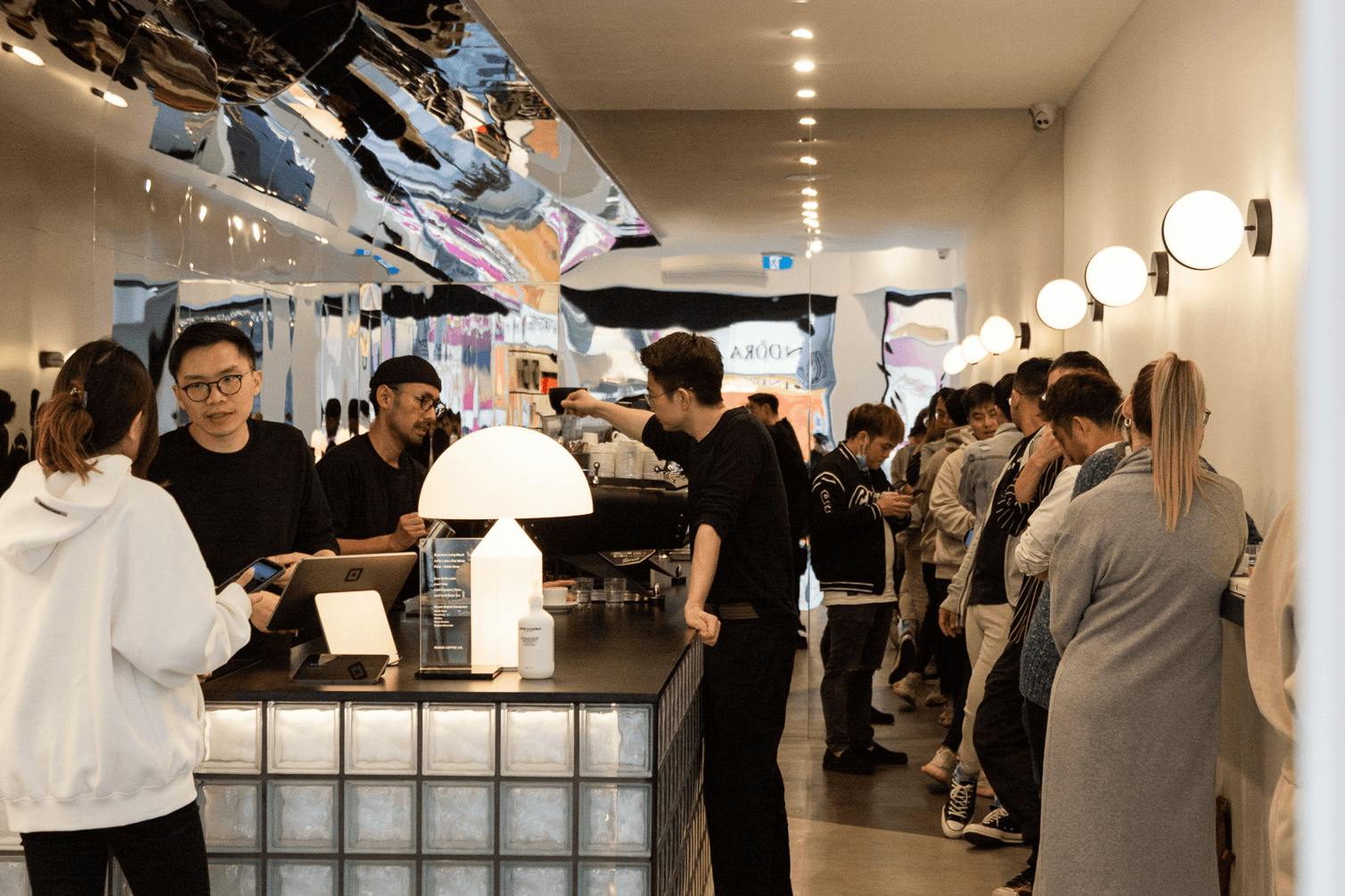 A busy cafe with people standing at a bar and a large overhead mirror.